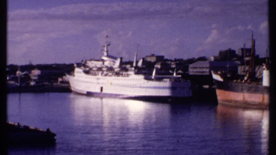 1969 Bahamas waterfront frame showing a white passenger vessel, low-rise harbor buildings, and a darker rust-colored vessel nearby.