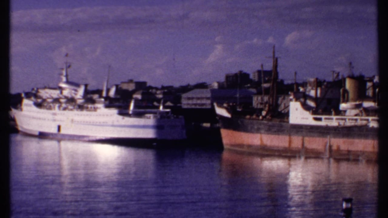 1969 Bahamas 8mm frame of a harbor waterfront with a white passenger vessel, nearby vessels, and low buildings along the shore.