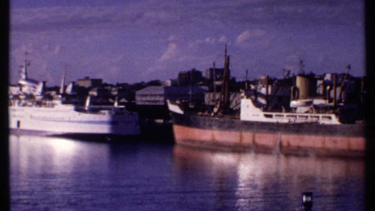 1969 Bahamas harbor frame showing a white vessel beside a rust-streaked cargo ship, with waterfront buildings and calm reflections.