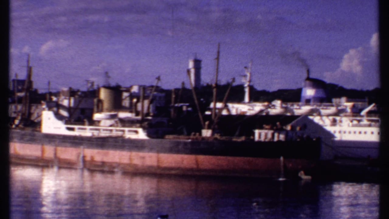 1969 Bahamas waterfront frame showing a white passenger vessel, a cargo ship, a water tower, and harbor structures along calm water.