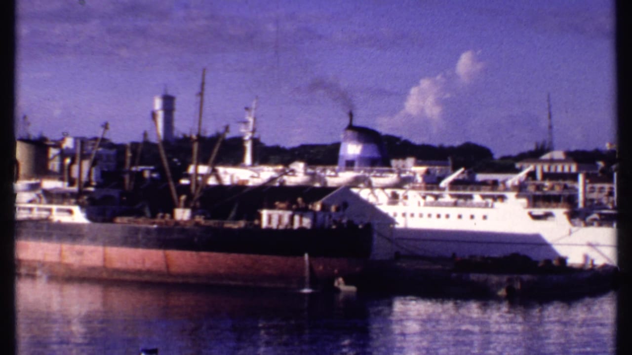 1969 Bahamas harbor frame with a white passenger ship behind a darker cargo vessel, plus a water tower, dock structures, and still water.