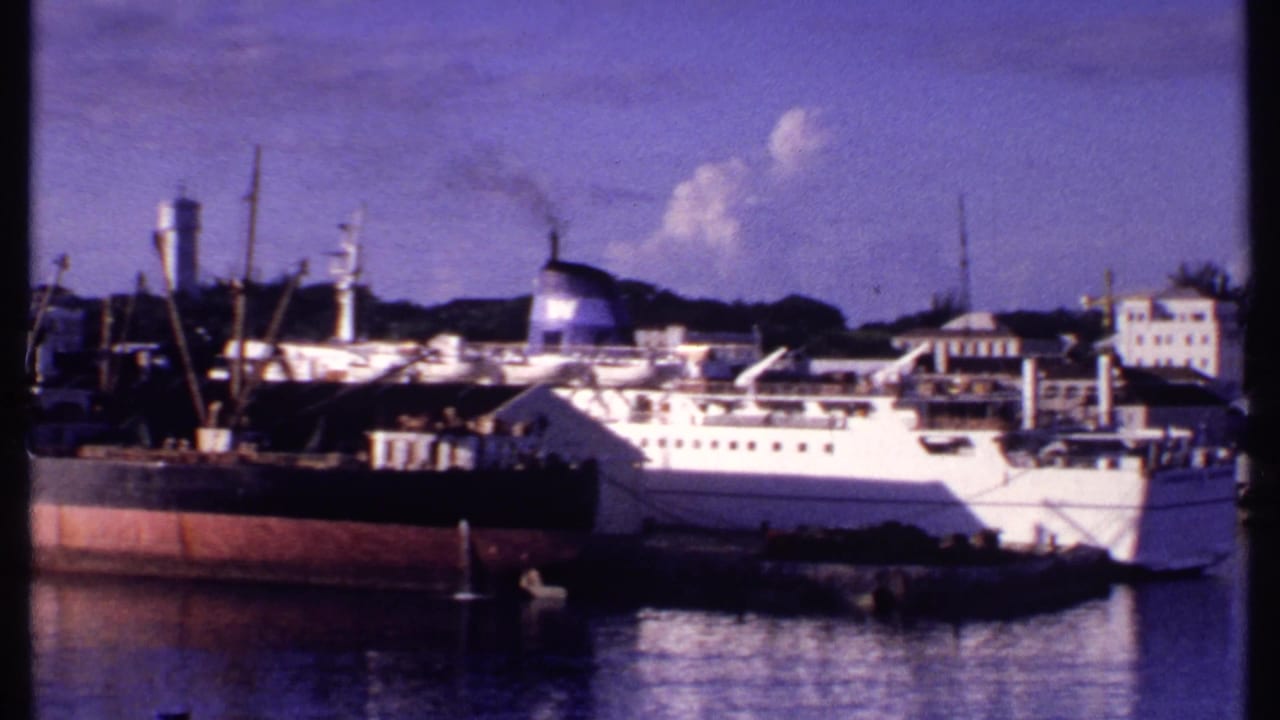 1969 Bahamas harbor view with a white passenger ship, darker cargo vessel, dock cranes, and waterfront buildings across calm water.
