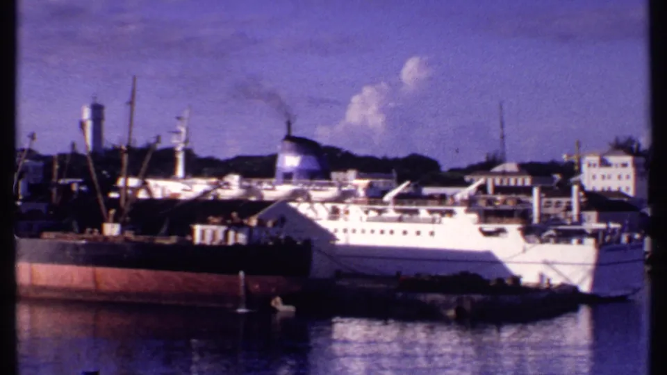 1969 Bahamas harbor view with a white passenger ship, darker cargo vessel, dock cranes, and waterfront buildings across calm water.