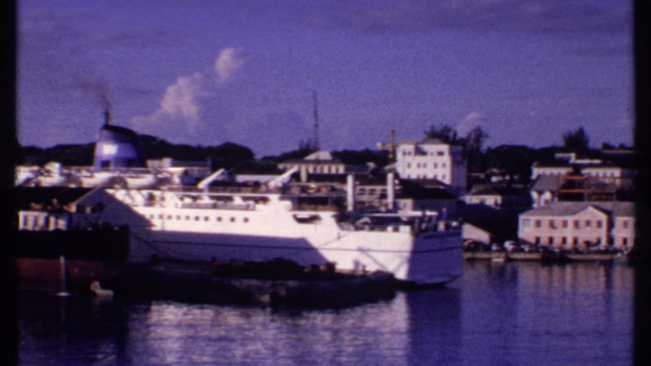 1969 Bahamas harbor frame showing the passenger ship alongside low waterfront buildings, dock equipment, and small boats.