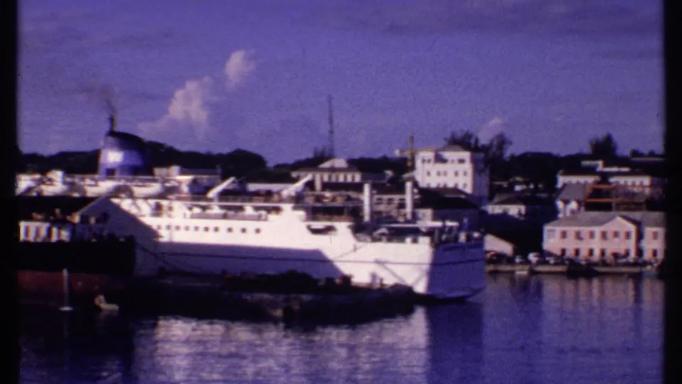 1969 Bahamas harbor frame showing the passenger ship alongside low waterfront buildings, dock equipment, and small boats.