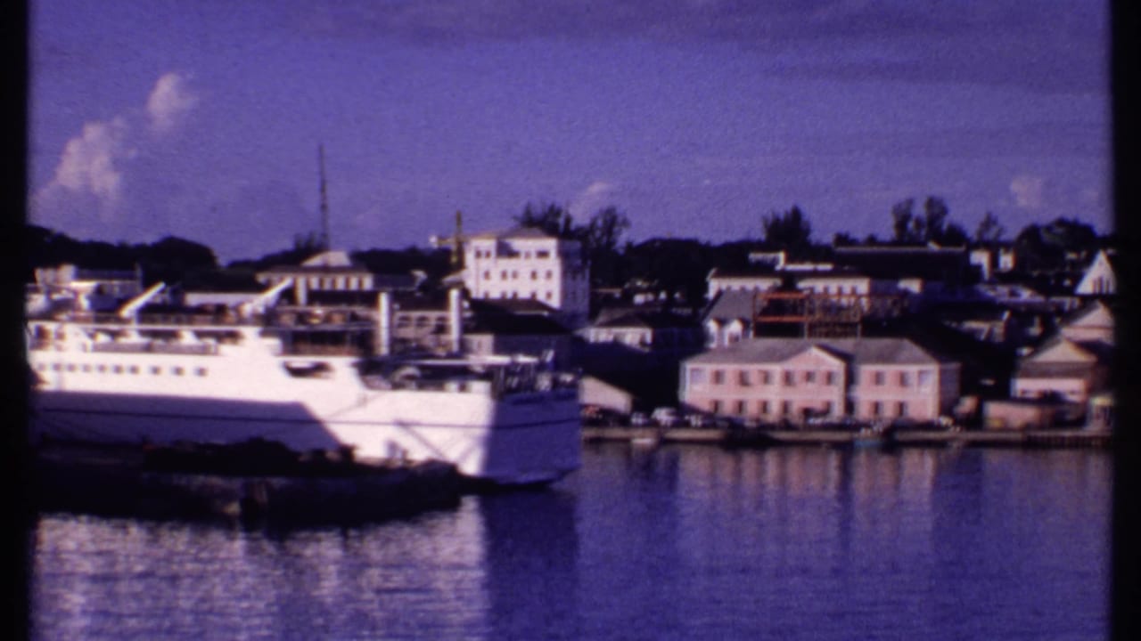 1969 Bahamas harbor pan with the passenger ship moving partly out of frame, revealing pastel waterfront buildings and calm water.
