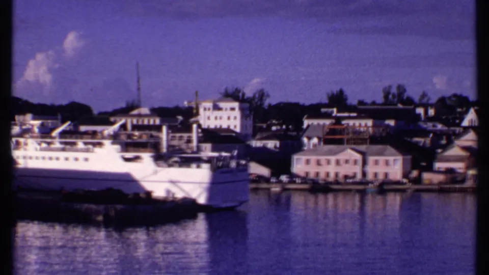 1969 Bahamas harbor pan with the passenger ship moving partly out of frame, revealing pastel waterfront buildings and calm water.