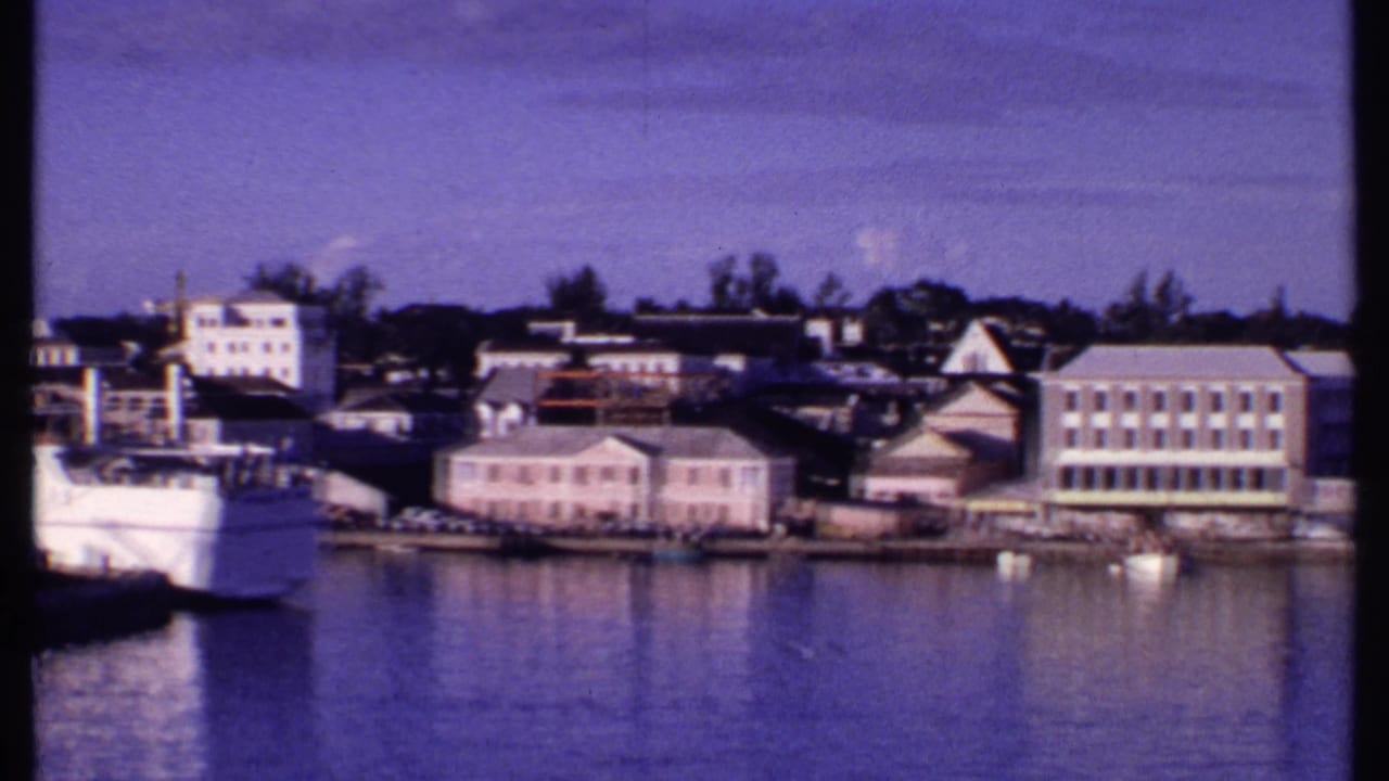 1969 Bahamas waterfront view with the bow of a passenger ship, pastel buildings, a larger block building, and still harbor water.