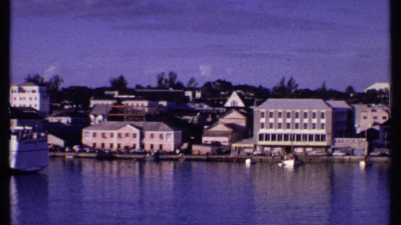 1969 Bahamas home-movie view of a waterfront town with pale buildings, moored boats, trees, and harbor reflections.