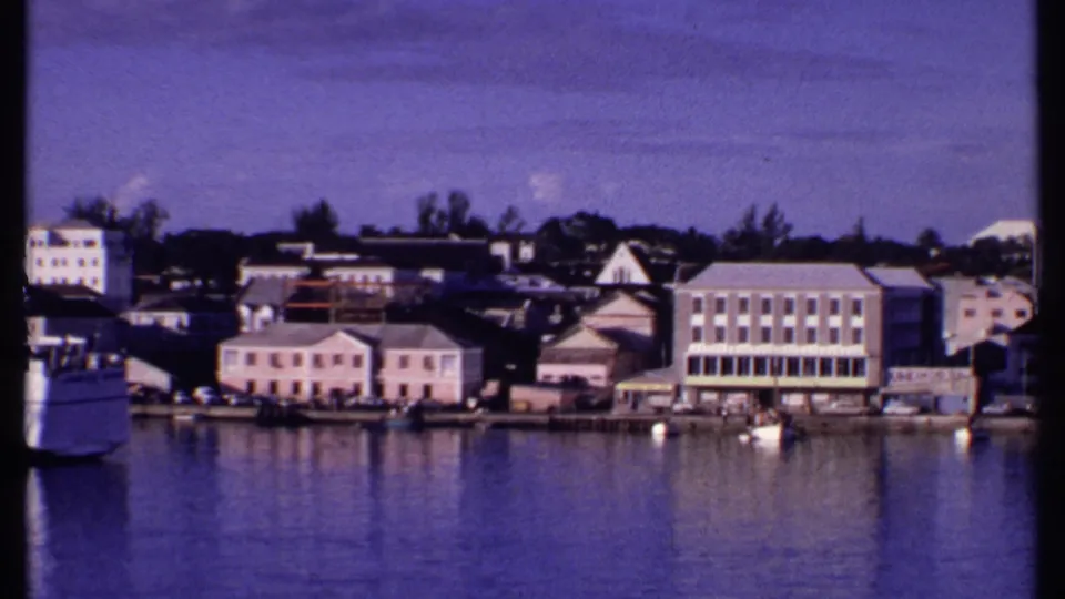 1969 Bahamas home-movie view of a waterfront town with pale buildings, moored boats, trees, and harbor reflections.