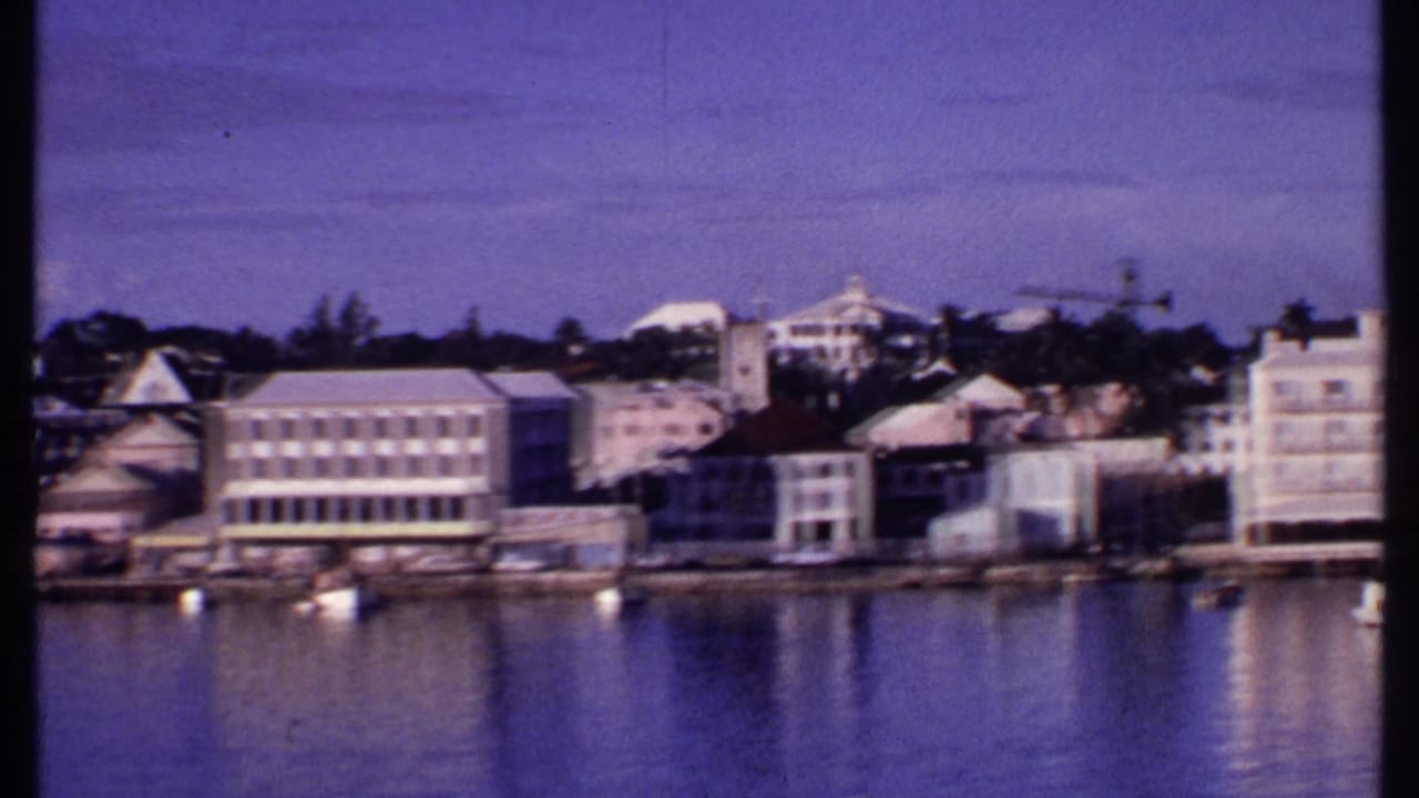 1969 Bahamas waterfront pan showing pale harbor buildings, small moored boats, trees, and soft reflections on the water.
