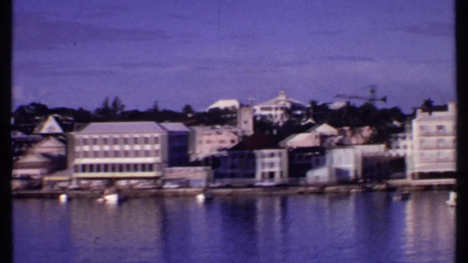 1969 Bahamas waterfront pan showing pale harbor buildings, small moored boats, trees, and soft reflections on the water.