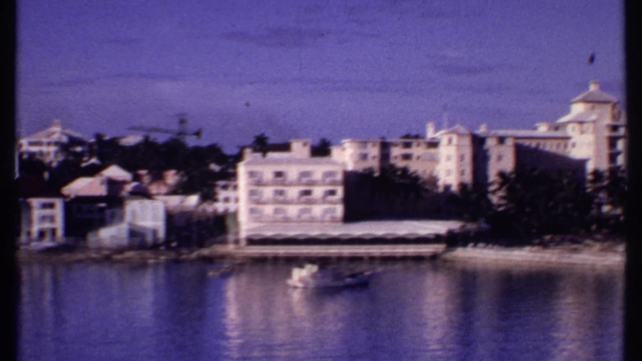 1969 Bahamas home-movie frame with larger waterfront buildings, a small boat on calm water, trees, and a purple-tinted sky.