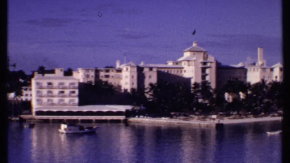 1969 Bahamas 8mm frame showing pale waterfront buildings, palms, and small boats across calm blue harbor water.