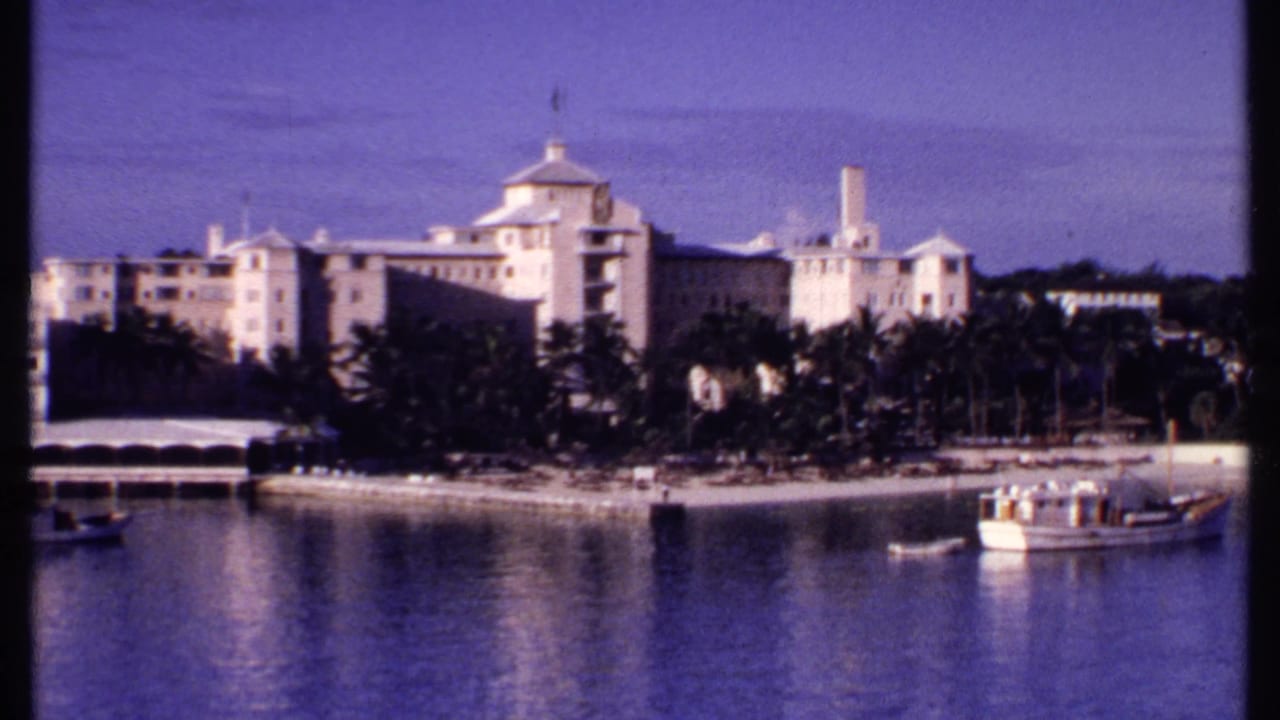 1969 Bahamas harbor view with a large pale waterfront complex, palm-lined shore, small boat, and larger vessel.
