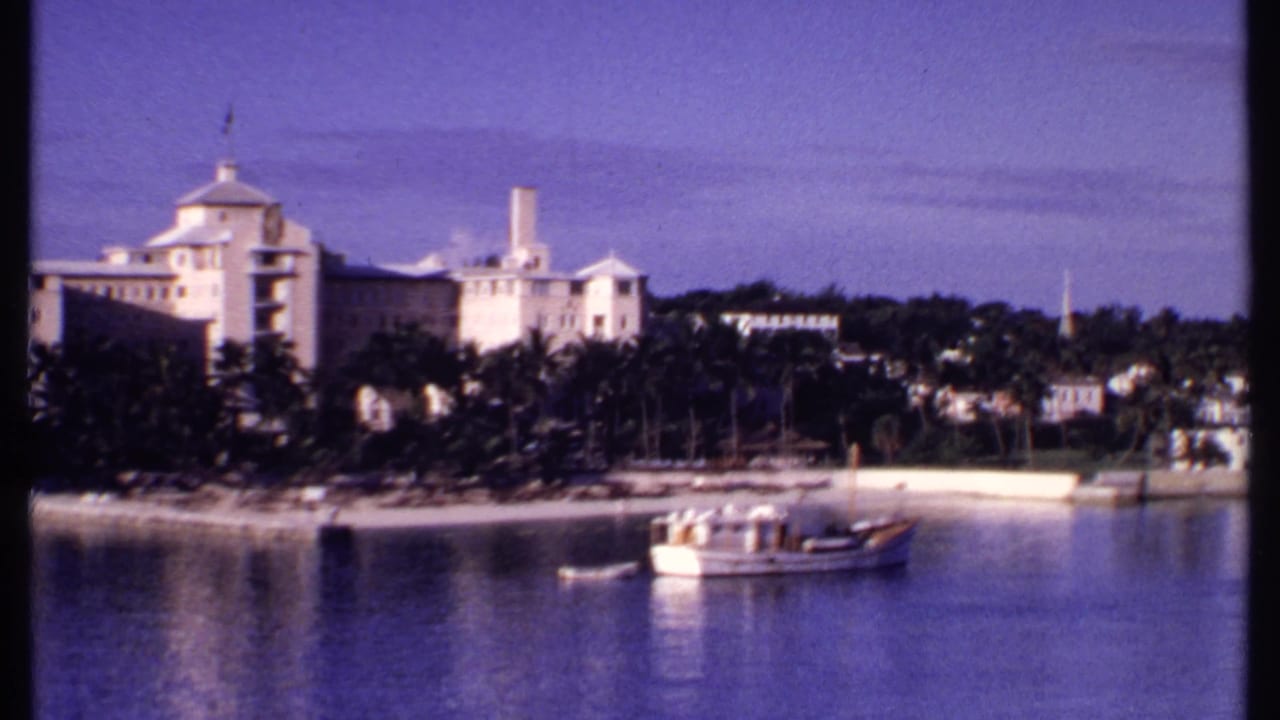 1969 Bahamas 8mm panorama frame showing waterfront buildings, palms, a sandy shore, and a larger boat on calm water.