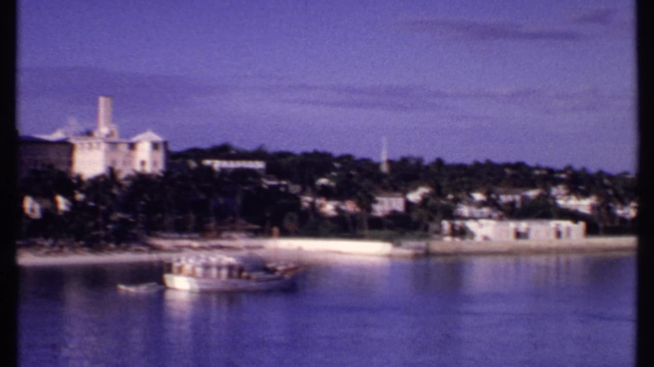1969 Bahamas harbor frame showing palms, pale waterfront buildings, distant hillside structures, and a larger boat on blue water.