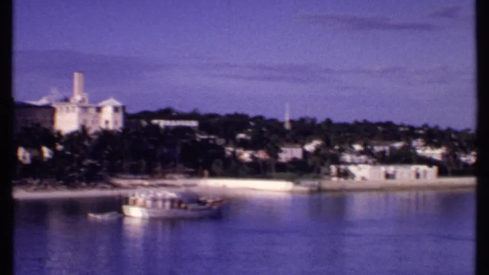 1969 Bahamas harbor frame showing palms, pale waterfront buildings, distant hillside structures, and a larger boat on blue water.