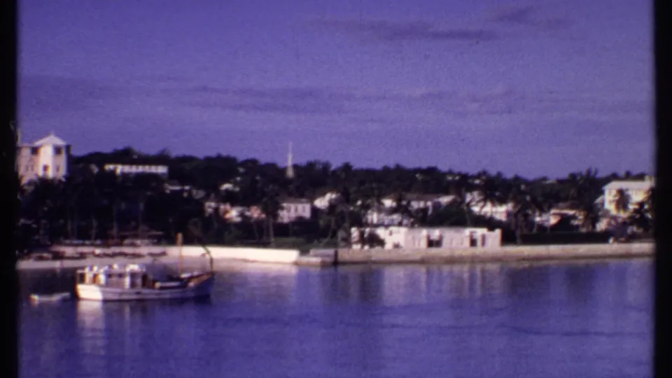 1969 Bahamas shoreline view with a small boat, calm water, palms, low white buildings, and a distant church steeple.
