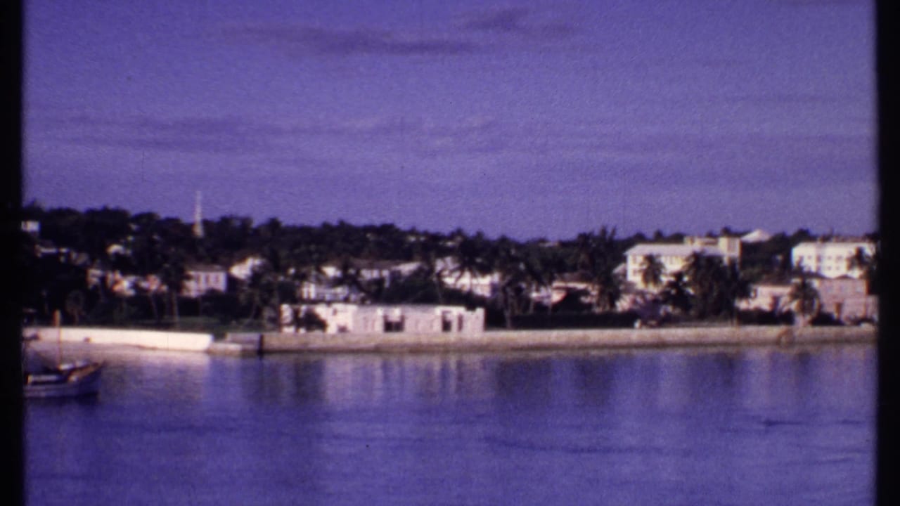 1969 Bahamas coastal view showing calm water, white buildings along shore, palms, and a small boat partly entering frame.