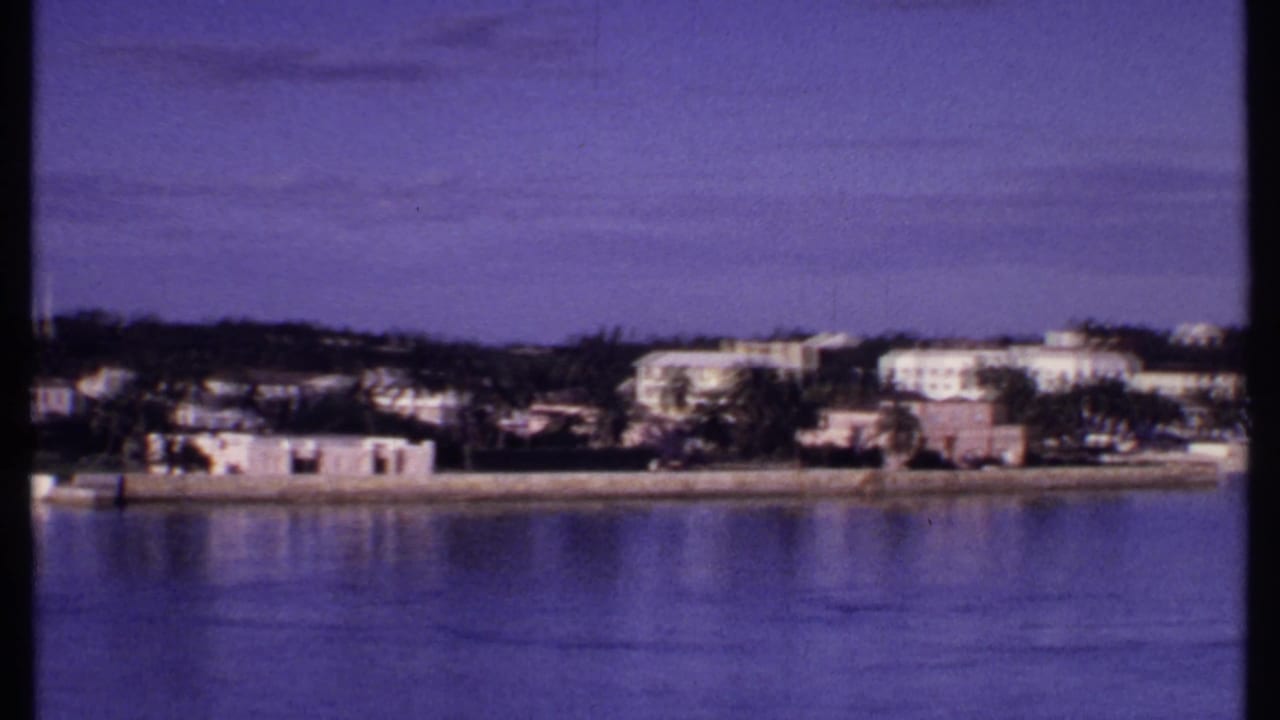 1969 Bahamas waterfront panorama with low coastal buildings, palms, calm reflective water, and open blue sky.