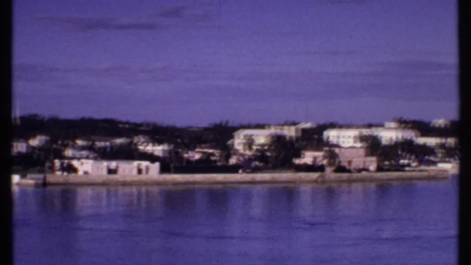1969 Bahamas waterfront panorama with low coastal buildings, palms, calm reflective water, and open blue sky.