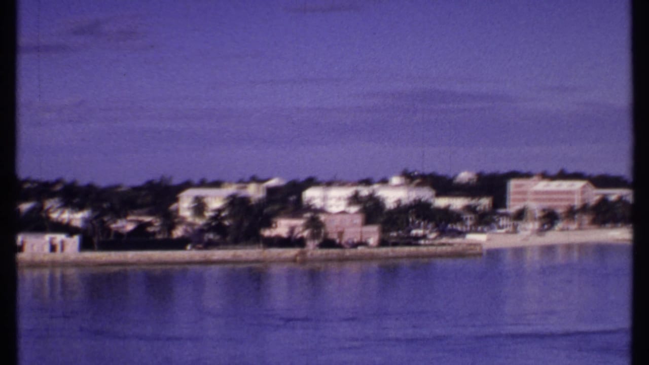 1969 Bahamas shoreline scene with calm water in the foreground and low white buildings among palms in the distance.