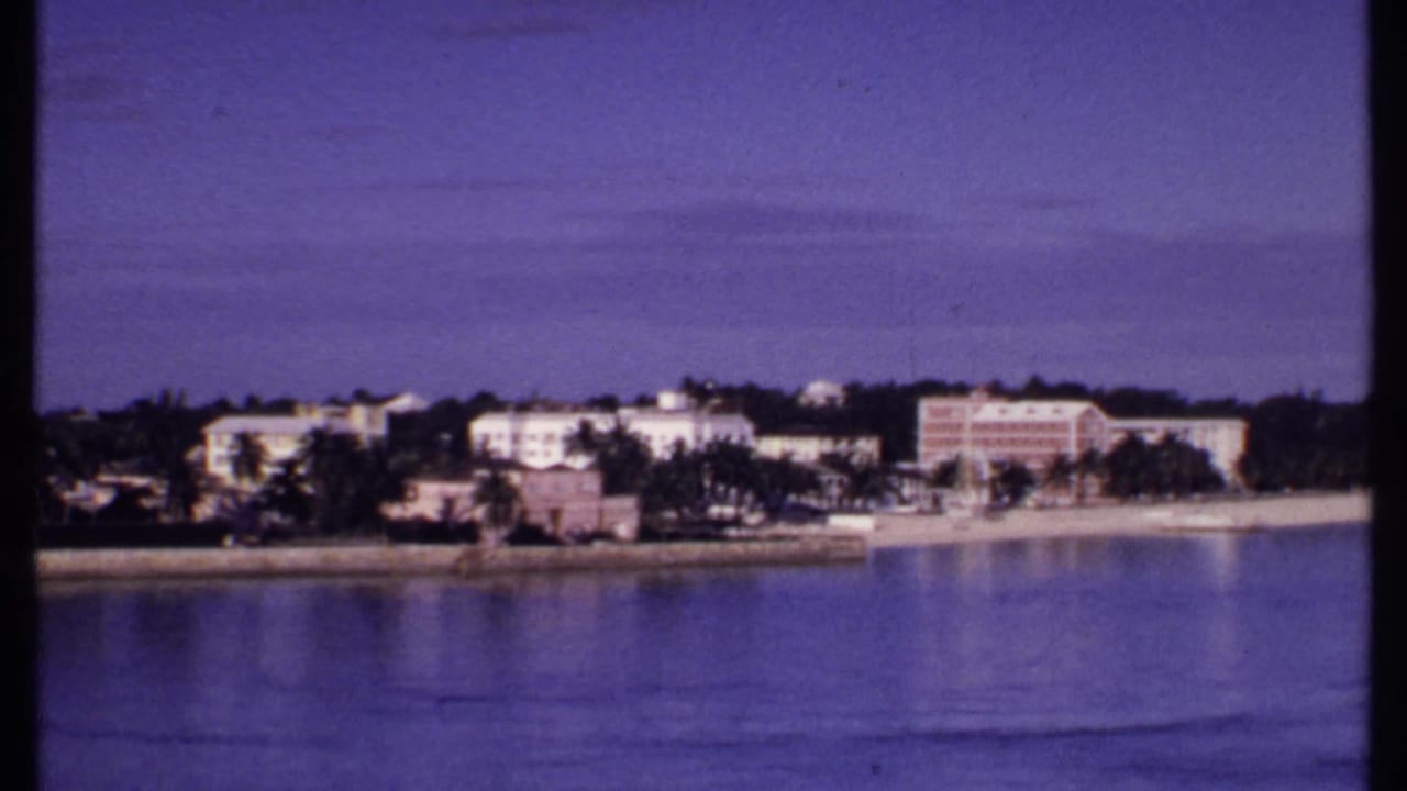 1969 Bahamas 8mm frame showing a resort-lined shoreline with palms, a sandy beach, and still blue water in the foreground.