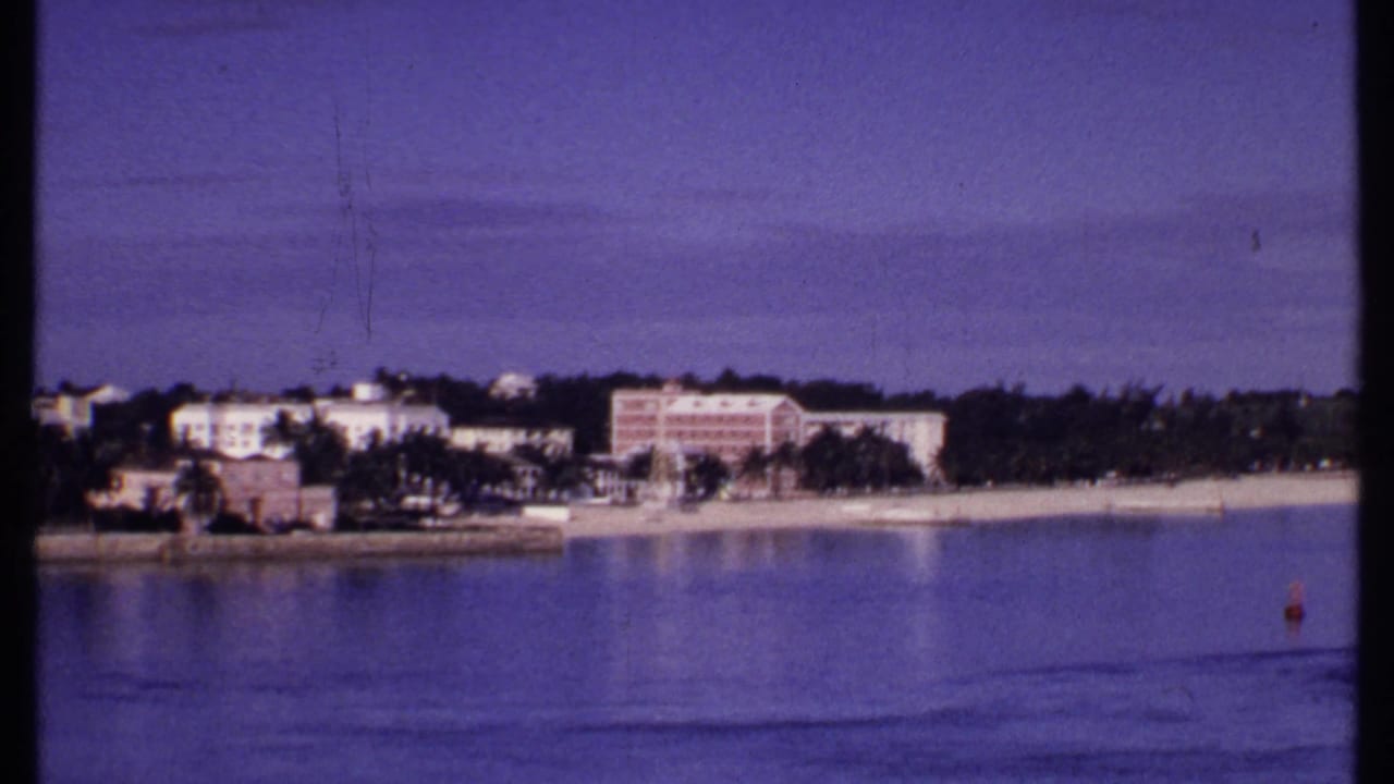 1969 Bahamas 8mm frame with pastel waterfront buildings, palm trees, sandy shore, calm ocean, and a small red buoy at right.