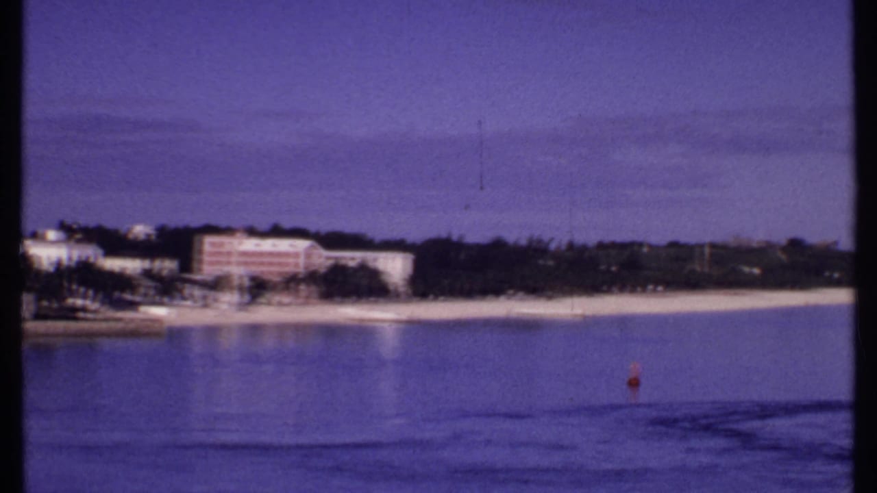 1969 Bahamas 8mm frame showing a distant beach resort shoreline, dark palms, calm water, and a red buoy near the right side.
