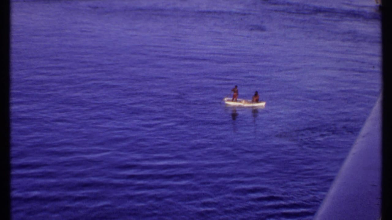 1969 Bahamas 8mm frame showing two distant people in a small open boat on calm blue water, with shoreline barely visible beyond.