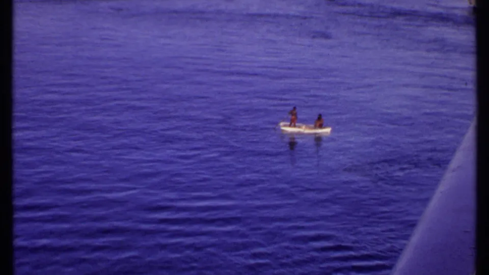 1969 Bahamas 8mm frame showing two distant people in a small open boat on calm blue water, with shoreline barely visible beyond.