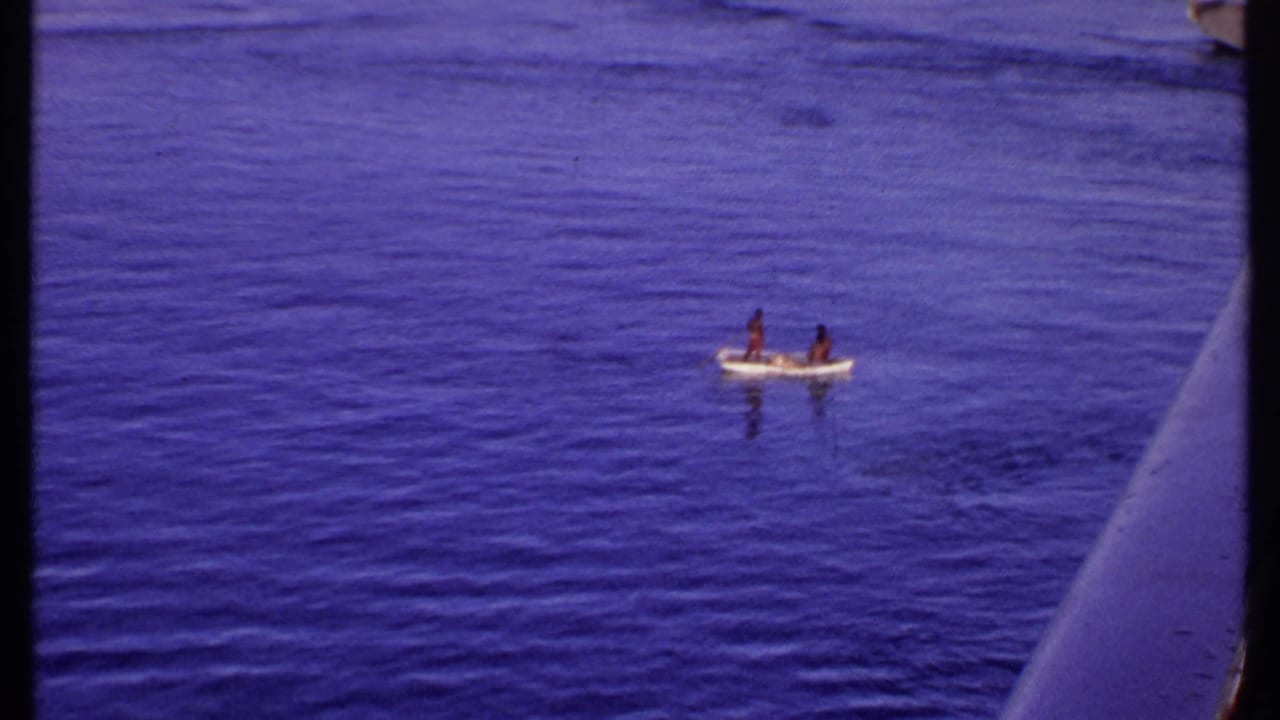 Small boat with two people on calm blue water beside a larger vessel.