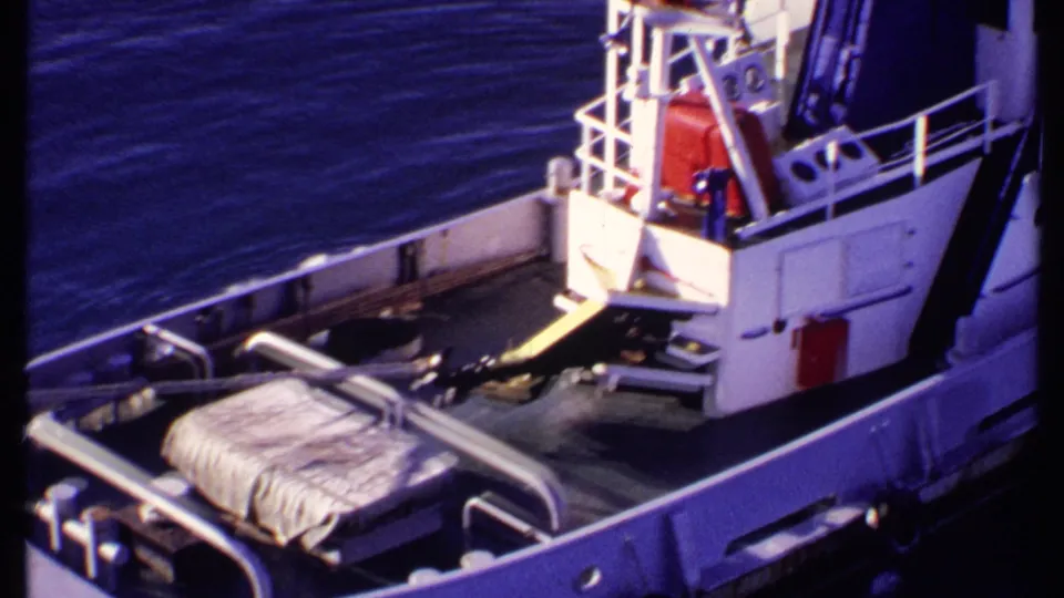 1969 Bahamas frame of a white vessel's deck and cabin area, with covered cargo and calm blue water nearby.