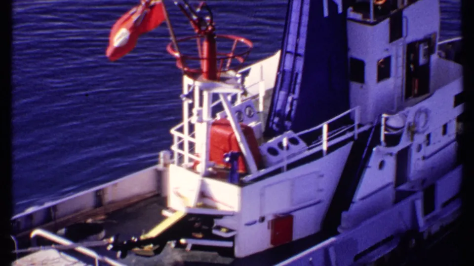 White vessel cabin and deck railings with a red flag, seen over blue water in 1969 Bahamas 8mm footage.