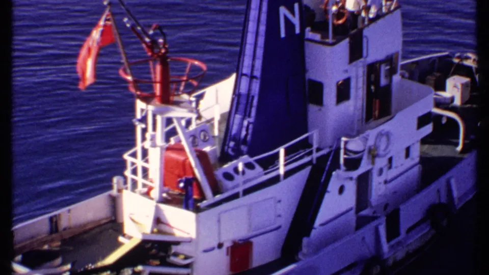1969 Bahamas 8mm frame showing a white and blue tugboat from above, with railings, a red flag, and a dark funnel marked N.