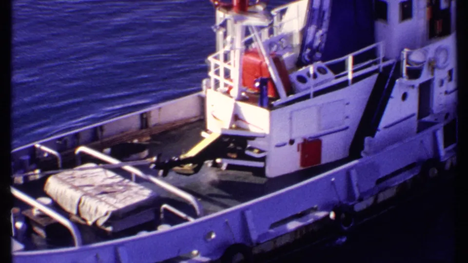 1969 Bahamas frame showing the tugboat's stern area and deck gear from above, with white railings, red flag, and surrounding calm blue water.