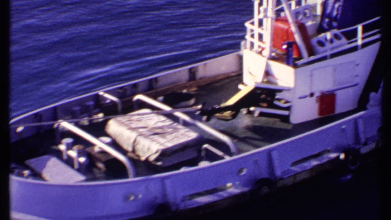 1969 Bahamas 8mm frame showing a small boat deck with metal railings, wrapped cargo, white cabin structure, and deep blue water.