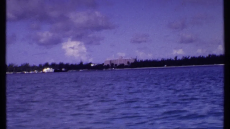 1969 Bahamas home-movie frame of a palm-lined coast and distant buildings across blue water beneath a cloudy sky.