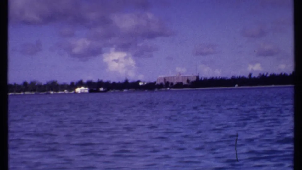 1969 Bahamas 8mm view across blue water toward a distant shoreline with palms, clouds, and a large resort-style building.