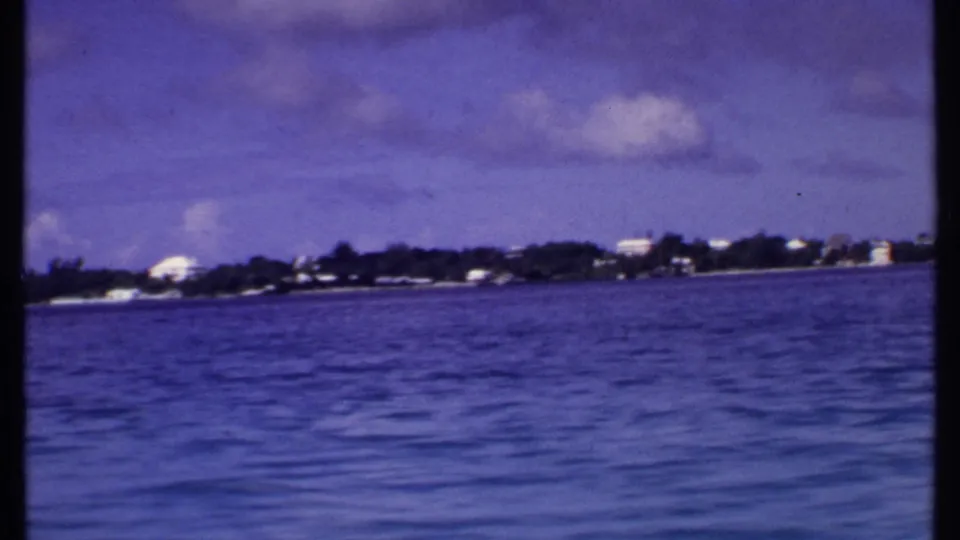 1969 Bahamas shoreline seen from a moving boat, with blue water in the foreground and low buildings along the distant coast.