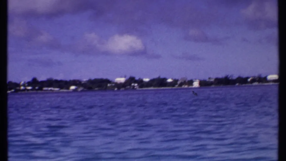 Vintage 8mm Bahamas travel frame with calm rippled water, dark shoreline vegetation, small distant structures, and a cloudy sky.