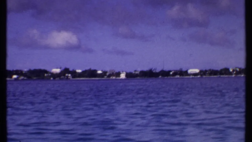 1969 Bahamas home-movie frame showing rippled blue water, a low tree-lined shore, scattered distant buildings, and soft 8mm color shift.