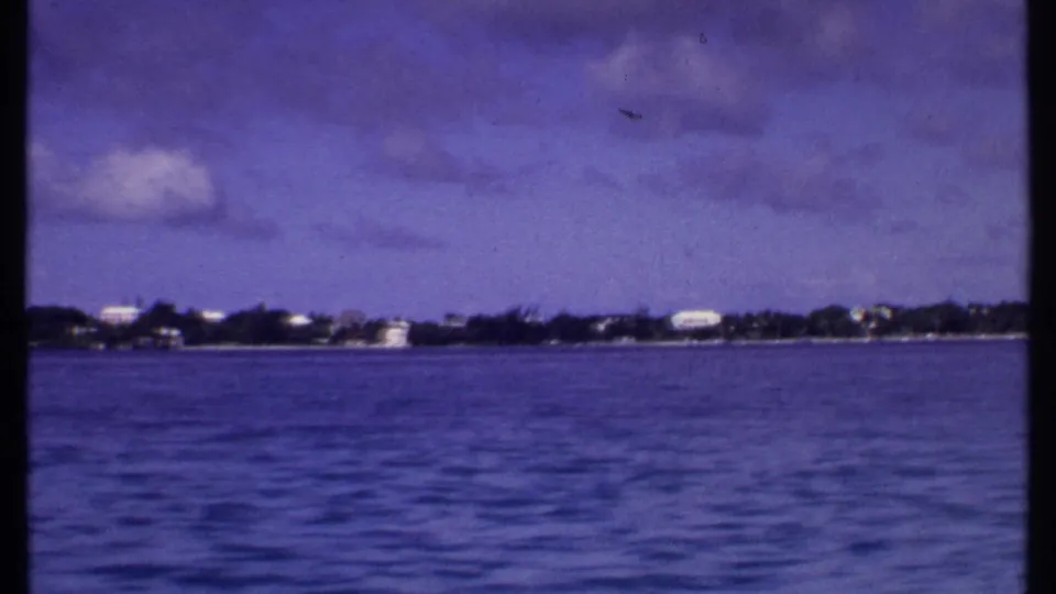 1969 Bahamas 8mm frame looking across blue water toward a distant tropical shoreline with small pale buildings under clouds.