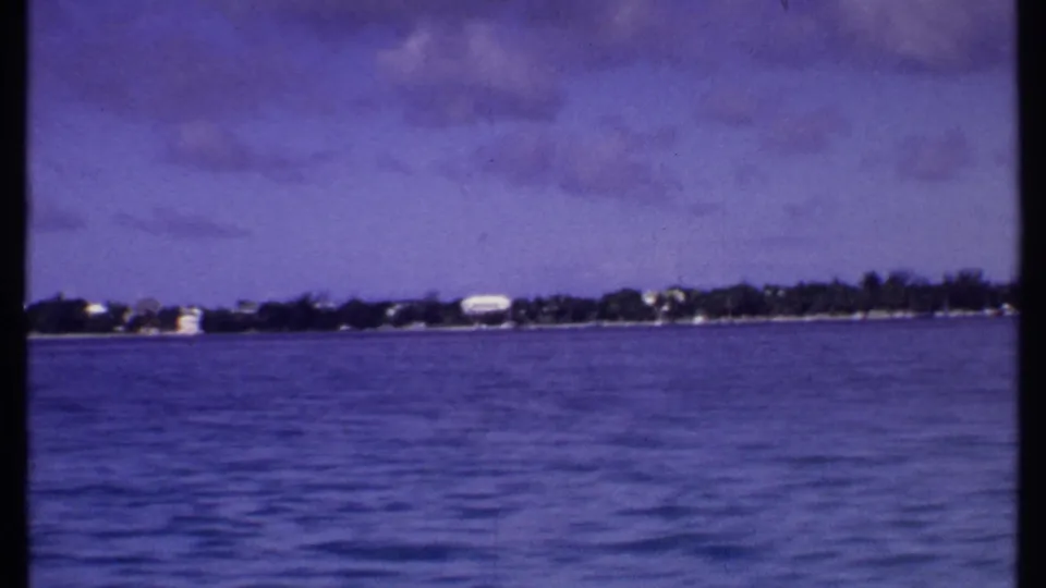 1969 Bahamas 8mm view over calm water toward a low coast with scattered white buildings and trees, split by the contact-sheet frame edge.