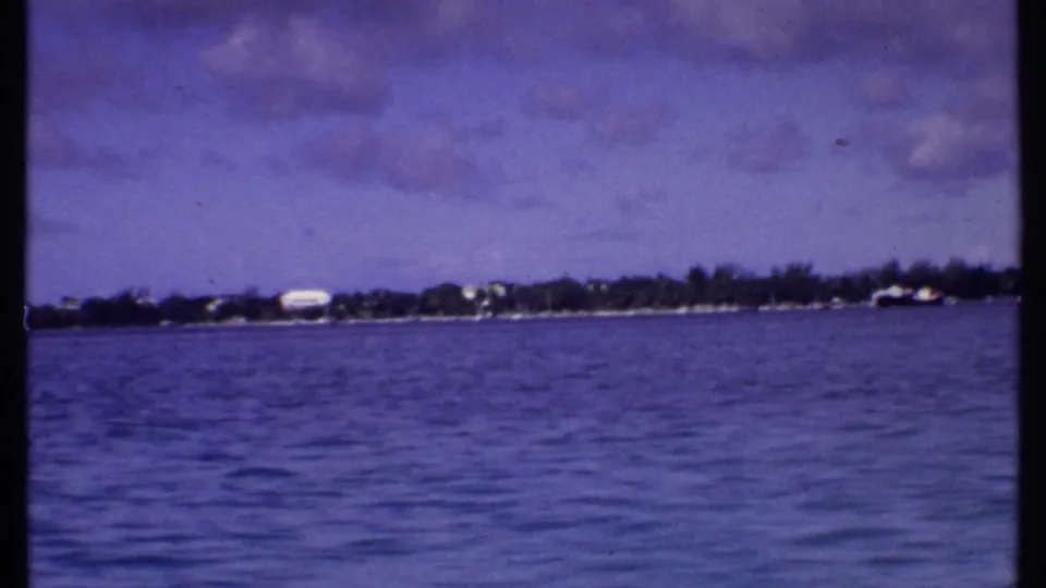 1969 Bahamas home-movie frame showing rippled blue water and a distant waterfront with low buildings and trees beneath a cloudy sky.