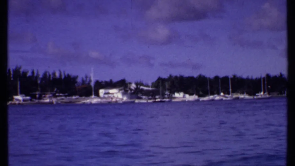 1969 Bahamas 8mm frame approaching a marina area with docked boats, sailboat masts, shoreline trees, and low waterfront buildings.