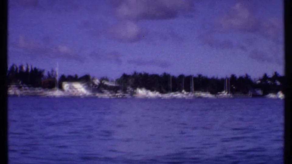 1969 Bahamas home-movie frame showing blue water, distant sailboats, palms, and low shoreline buildings under a purple-tinged sky.