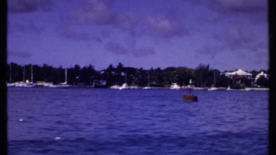 1969 Bahamas harbor frame with sailboats near shore, white waterfront buildings, palms, and a small red buoy or boat in the foreground water.