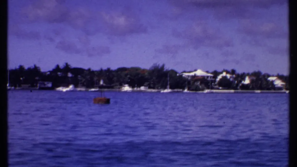 1969 Bahamas home-movie frame showing a tropical shoreline, moored sailboats, pale waterfront buildings, and a small red buoy or boat.